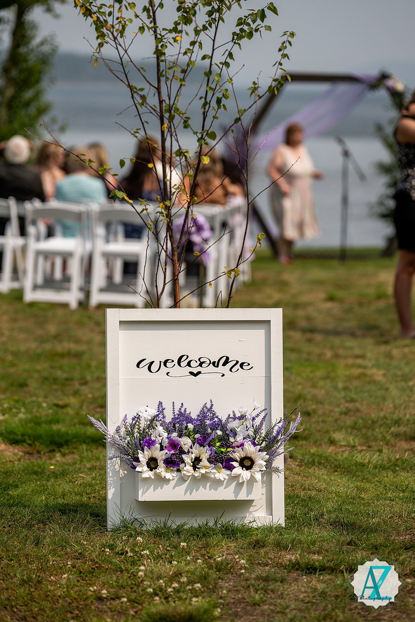 White welcome sign with purple and white flowers on outdoor lawn at waterfront wedding ceremony setup.