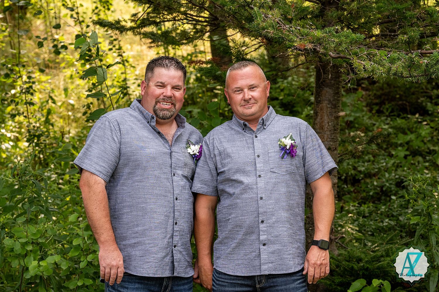Two people wearing matching grey button-down shirts pose together in a sunlit wooded outdoor setting.