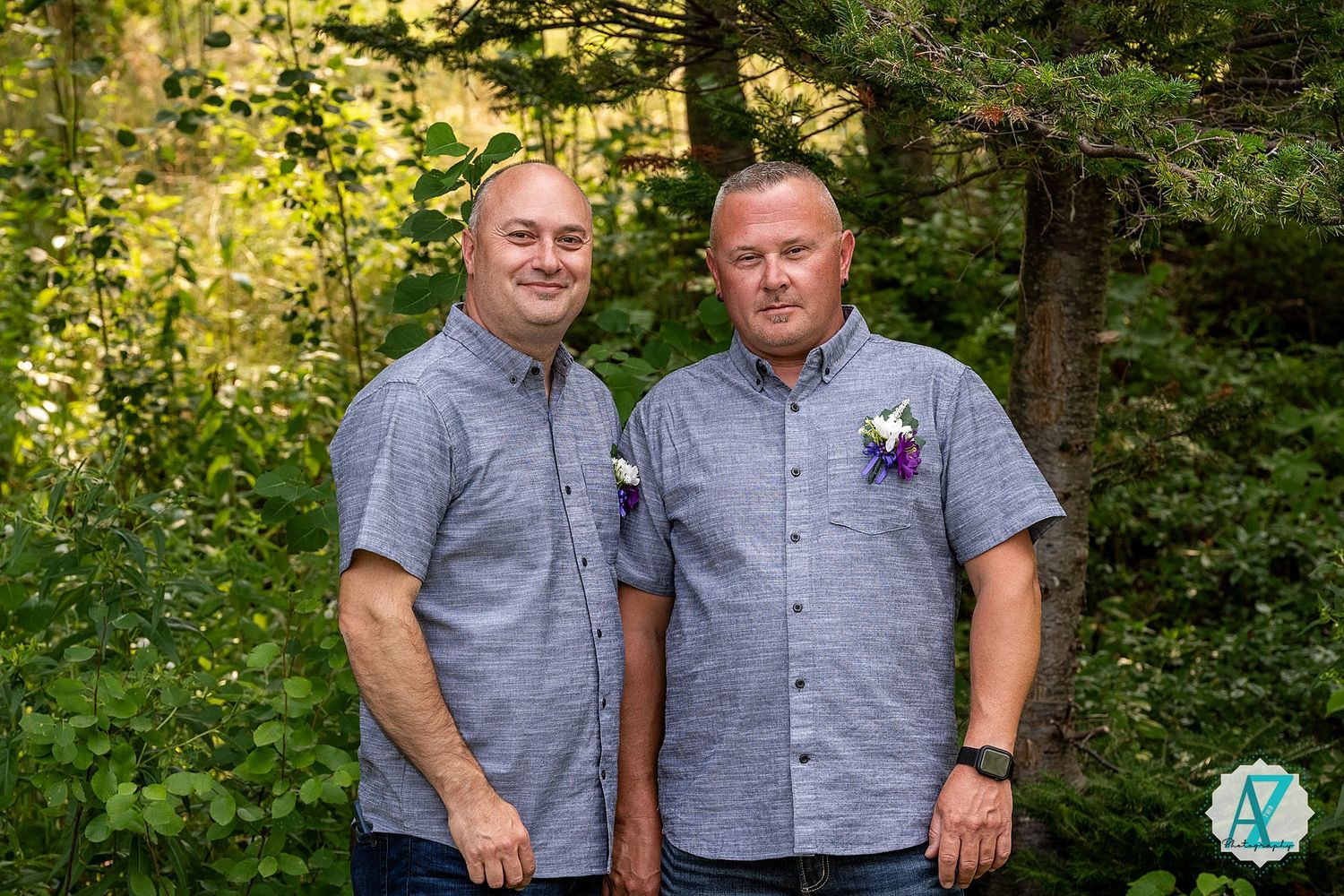 Two guests wearing matching gray button-down shirts pose together outdoors with boutonnieres in a wooded setting.