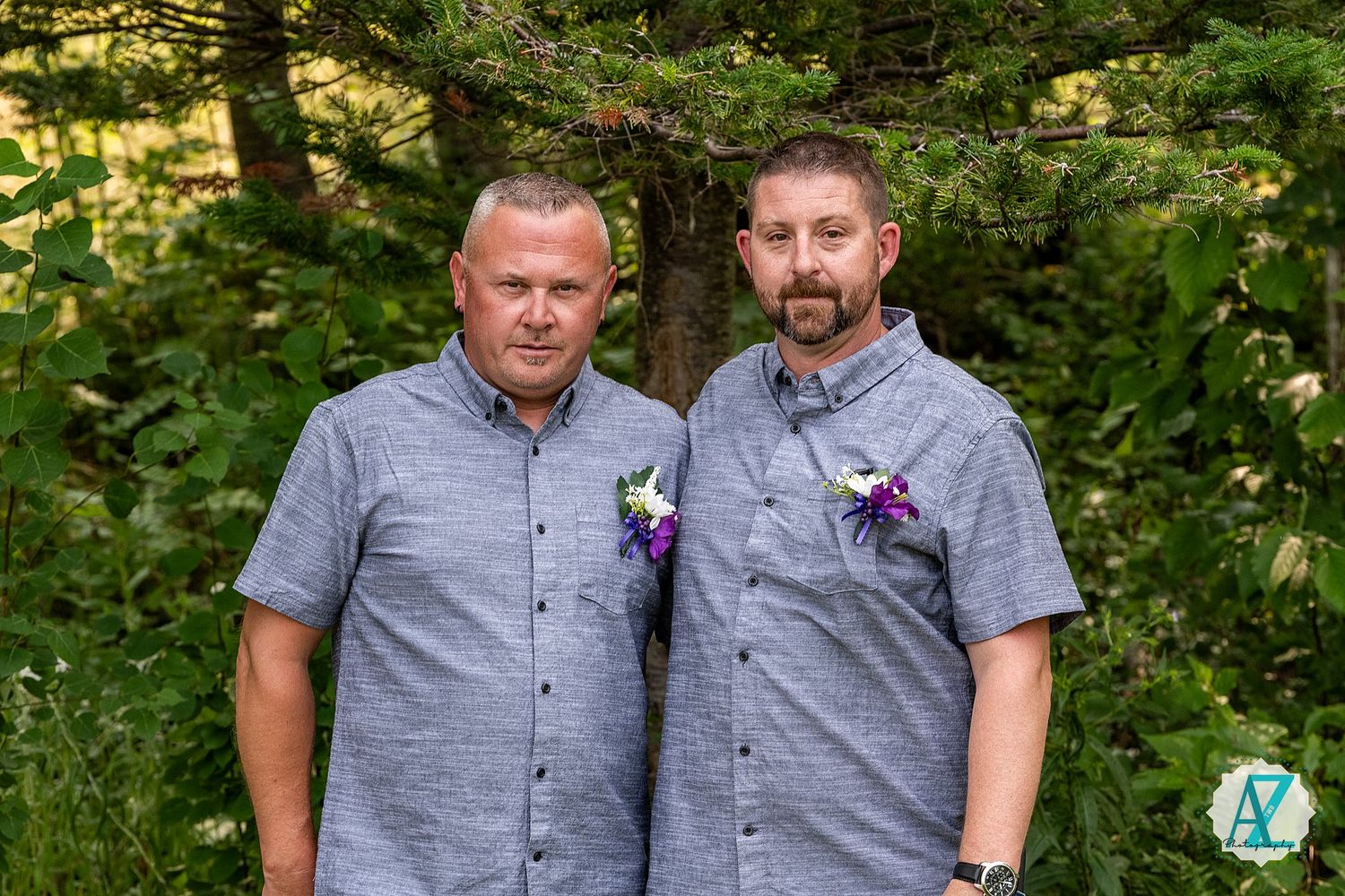 Two wedding attendants in coordinated gray shirts with purple boutonnieres stand together against a forest backdrop.