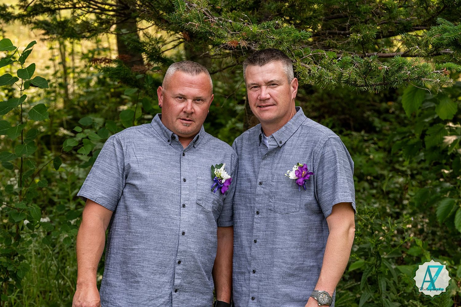 Two people in matching gray shirts stand together in a lush green forest setting with purple flowers on their lapels.