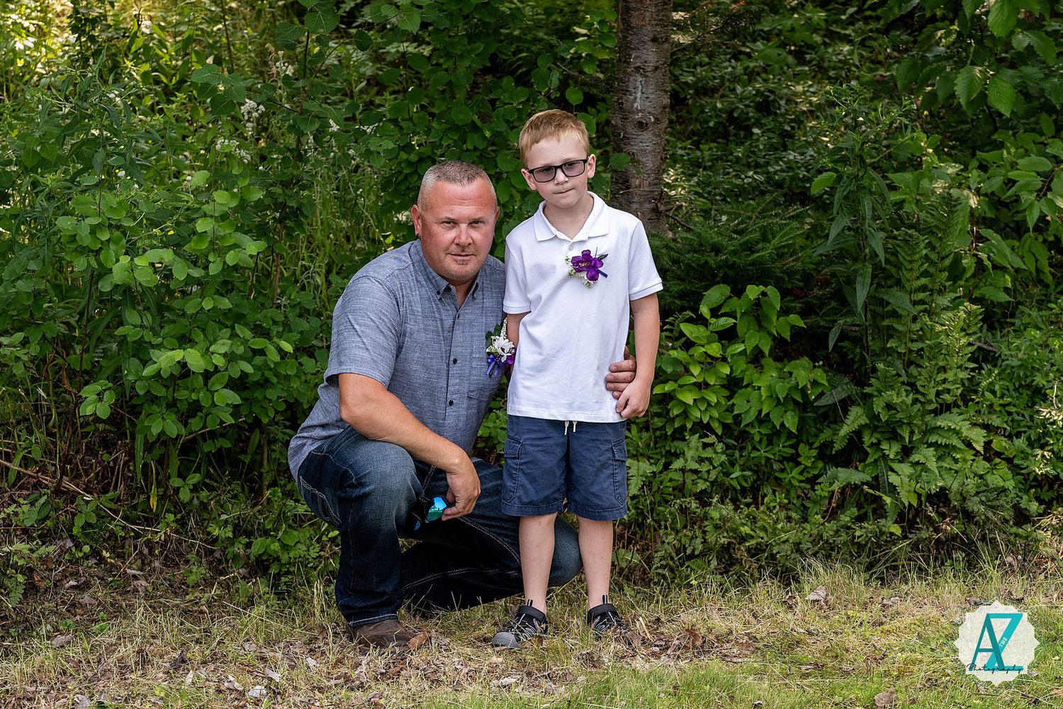 Two people pose together in front of lush green foliage on a sunny day outdoors.