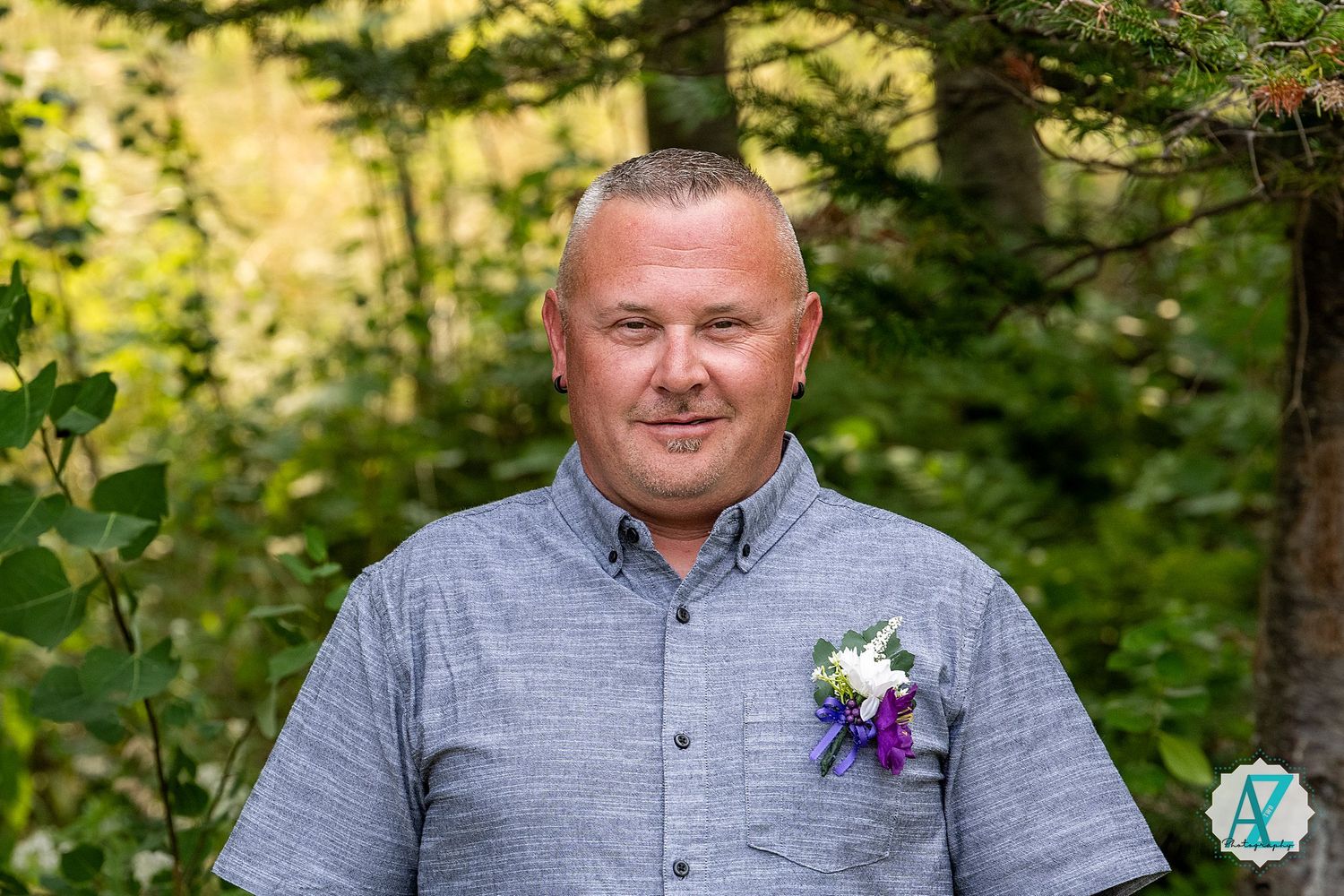 A person in a blue button-down shirt and purple boutonniere poses against a green forest background for a portrait.