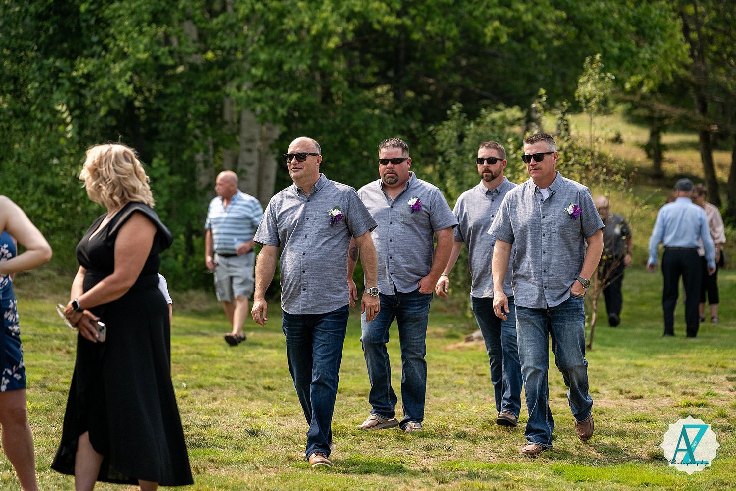 A group of casually dressed guests walk across a grassy outdoor area during an event on a sunny day.