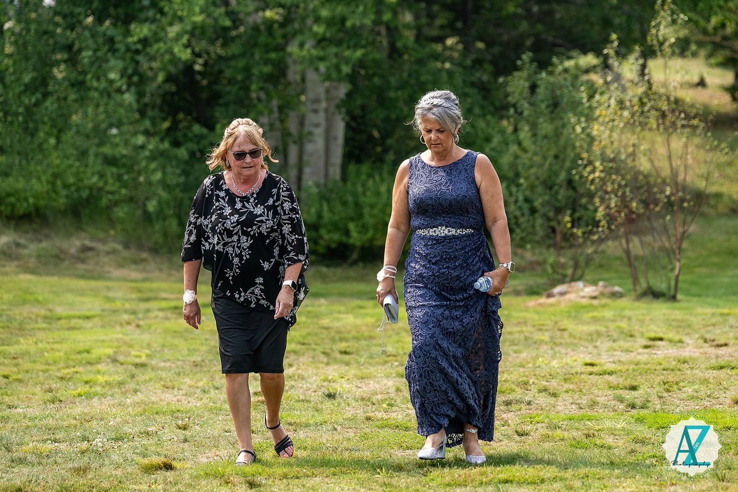 Two women walking together on grass with trees in the background during a sunny day.