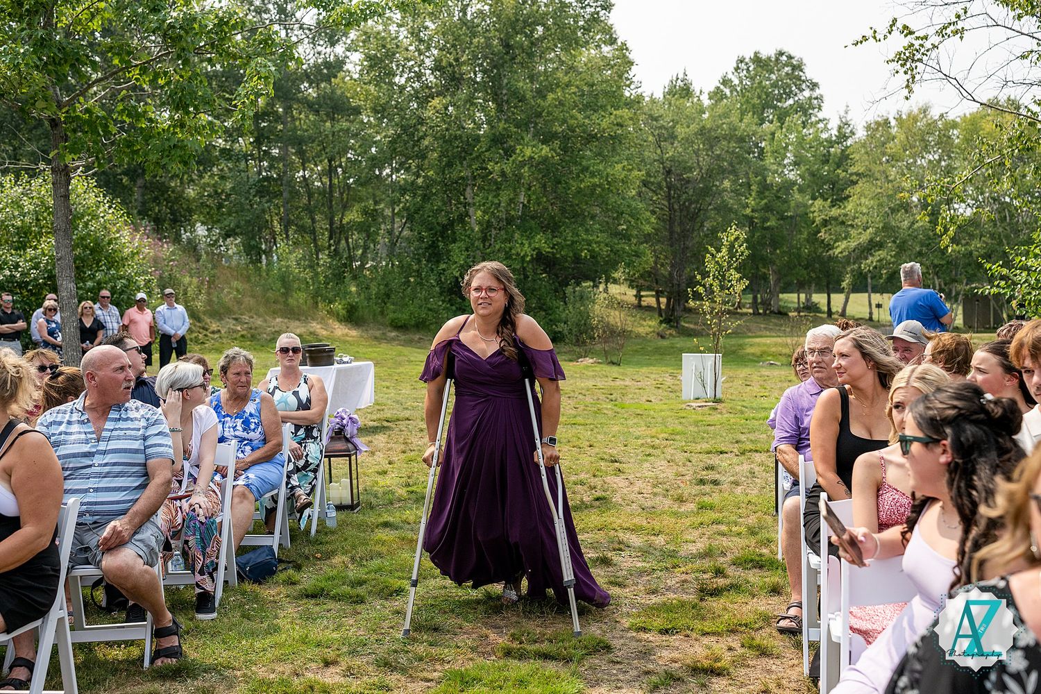A purple-dressed person walks down the aisle at an outdoor wedding ceremony with guests seated on white chairs.
