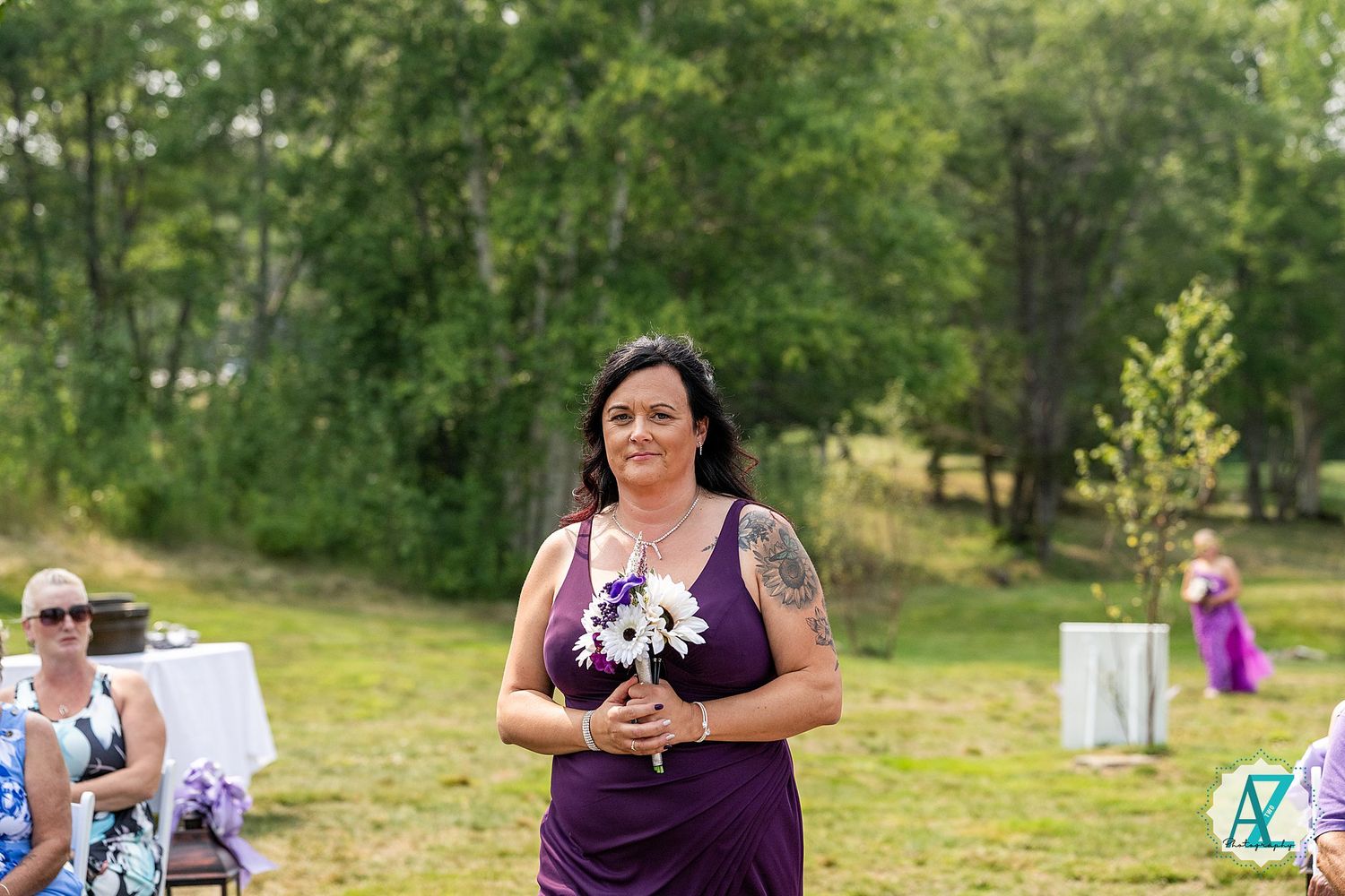 Outdoor wedding celebration with people gathered in sun-dappled woods wearing purple and white attire.