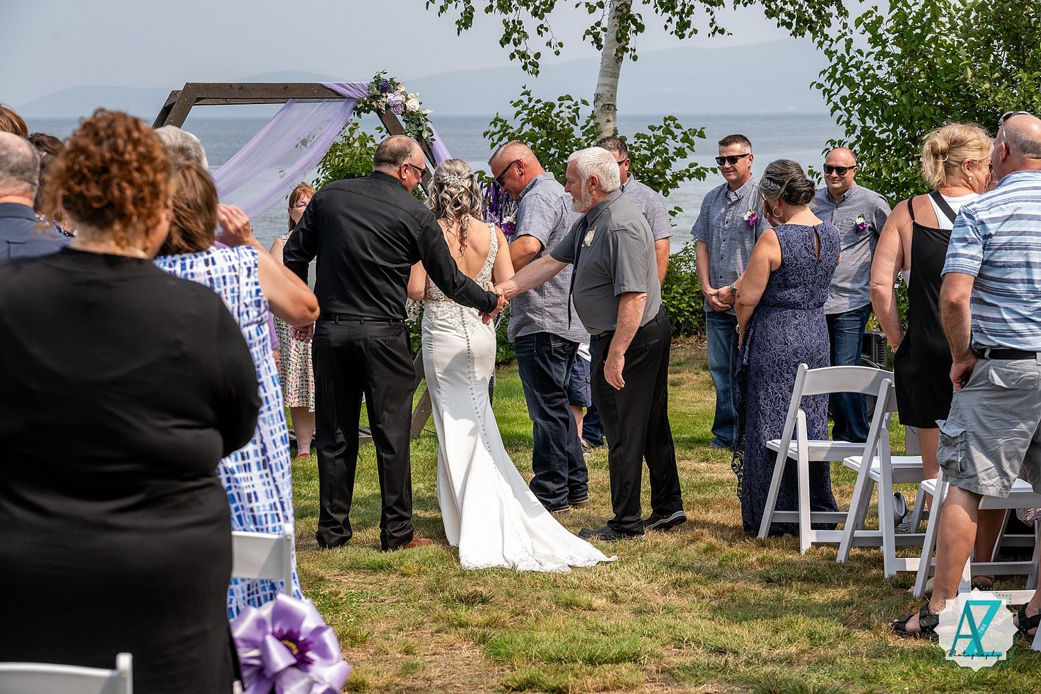Intimate outdoor wedding ceremony takes place on a grassy hillside with guests gathered around in casual summer attire.