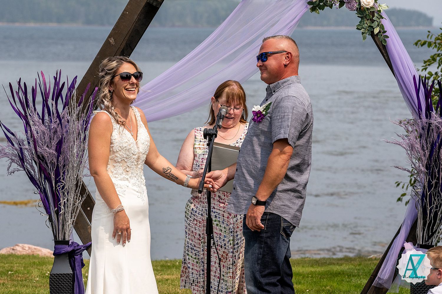 A couple exchanges vows during a lakeside wedding ceremony with purple decorations and scenic water views in the background.