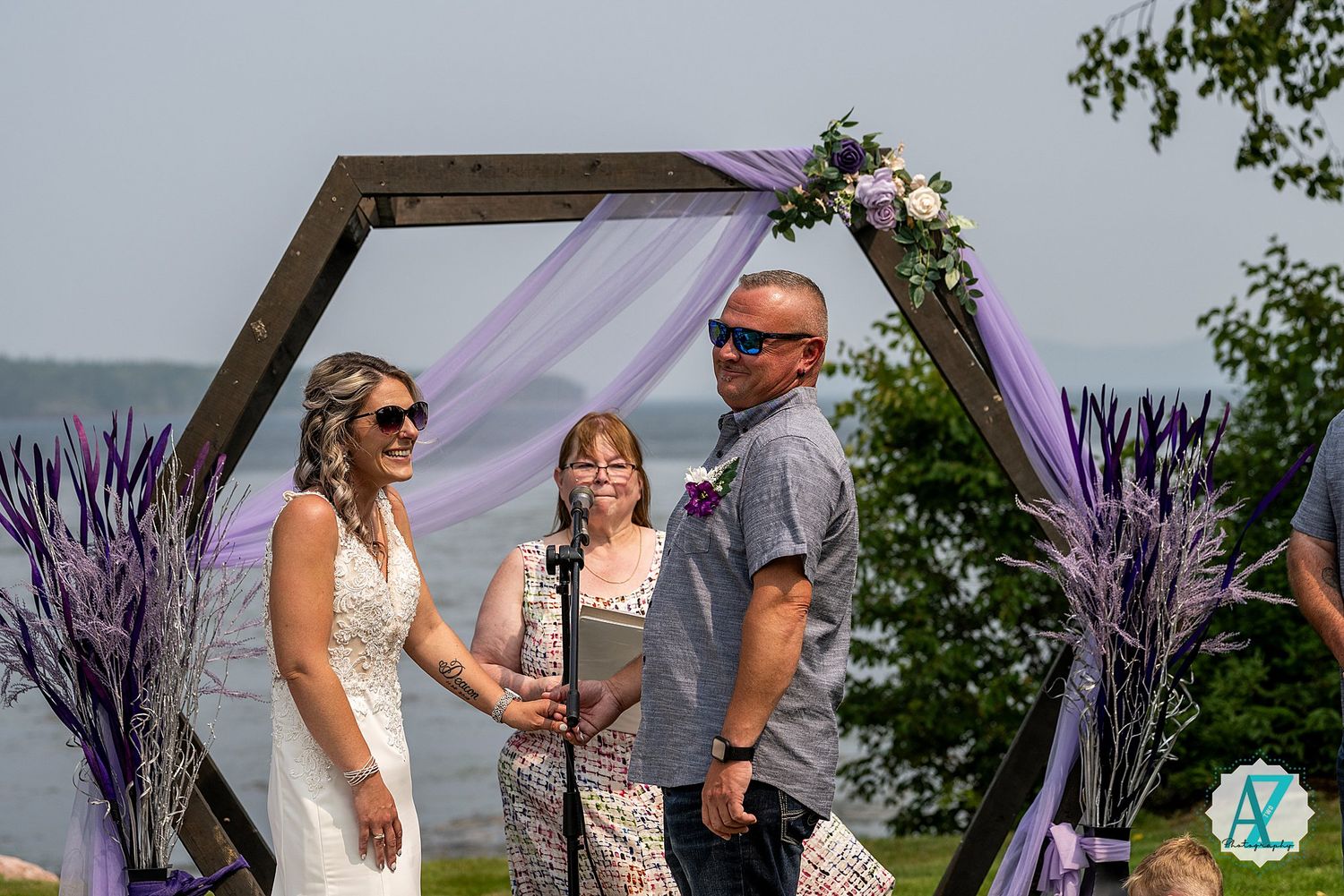 Outdoor wedding ceremony under a wooden arch decorated with purple fabric and flowers by the ocean.