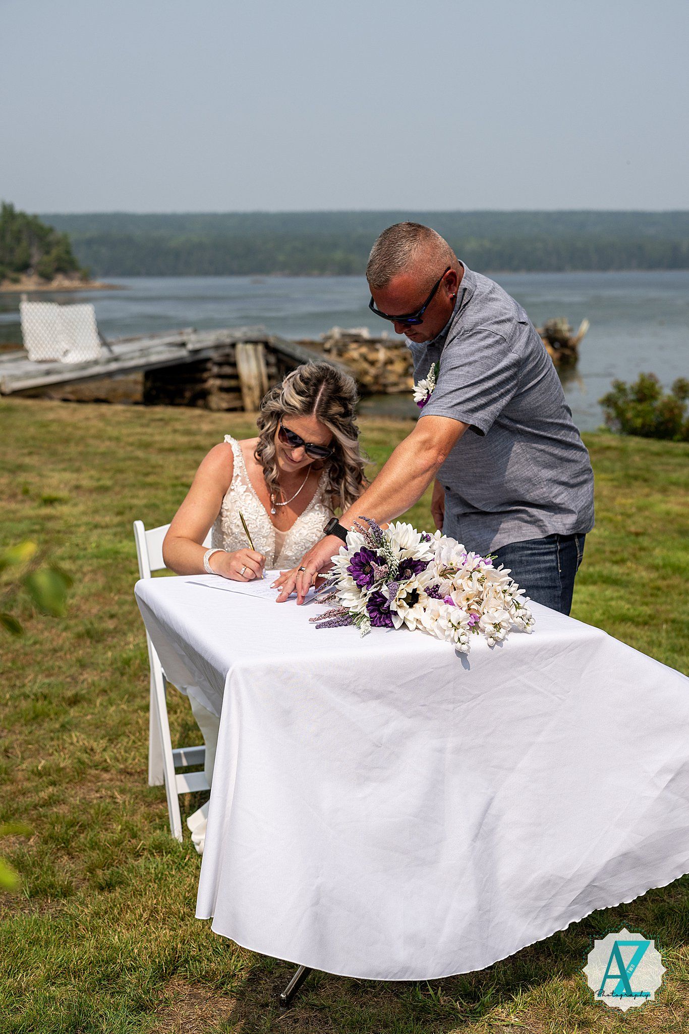 Wedding couple signing documents at a white table on a scenic oceanfront cliff.