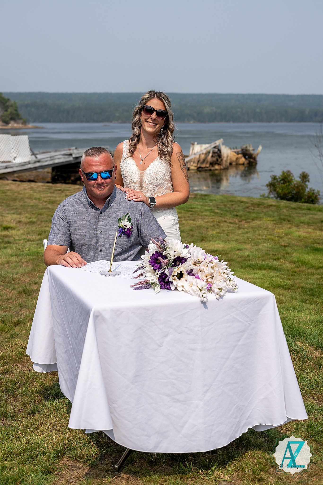 Couple signing marriage documents at a seaside outdoor ceremony with white tablecloth and purple flowers.
