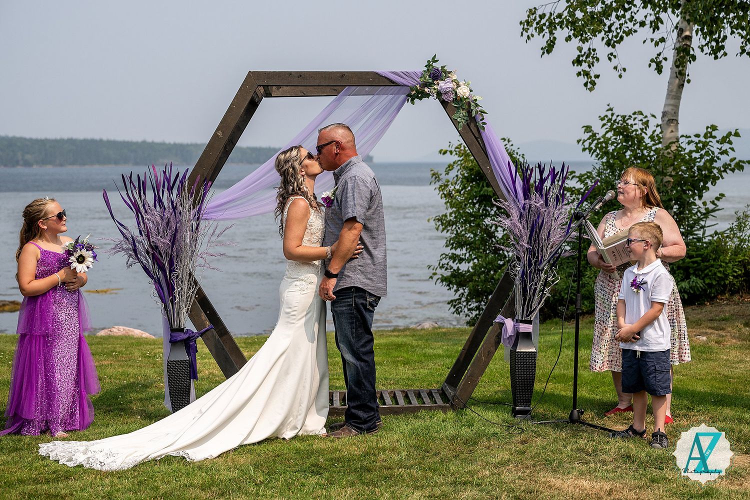Outdoor wedding ceremony with purple decor features hexagonal arch and lakeside backdrop.