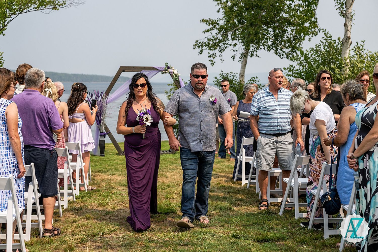 Wedding guests line white chairs as attendants walk down aisle during outdoor ceremony by water.