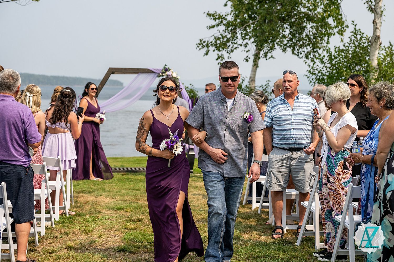 Wedding party members walk down aisle between rows of seated guests at outdoor lakeside ceremony.