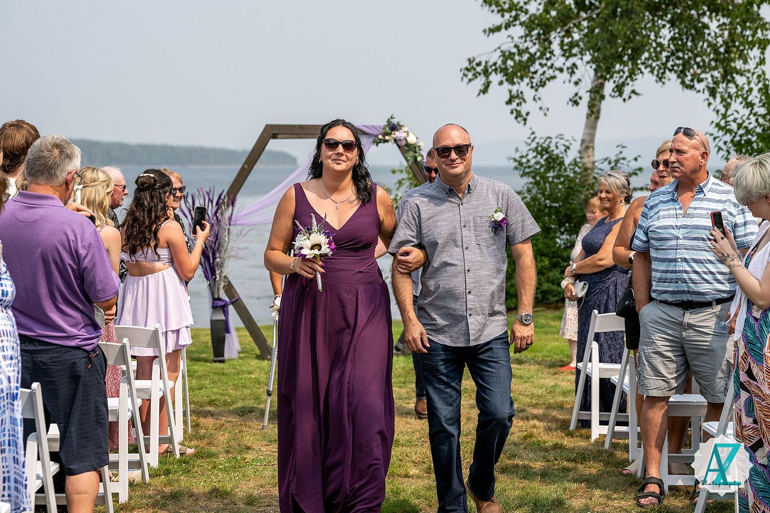 Wedding guests gather on grassy lakeside lawn near wooden arch during outdoor ceremony celebration.