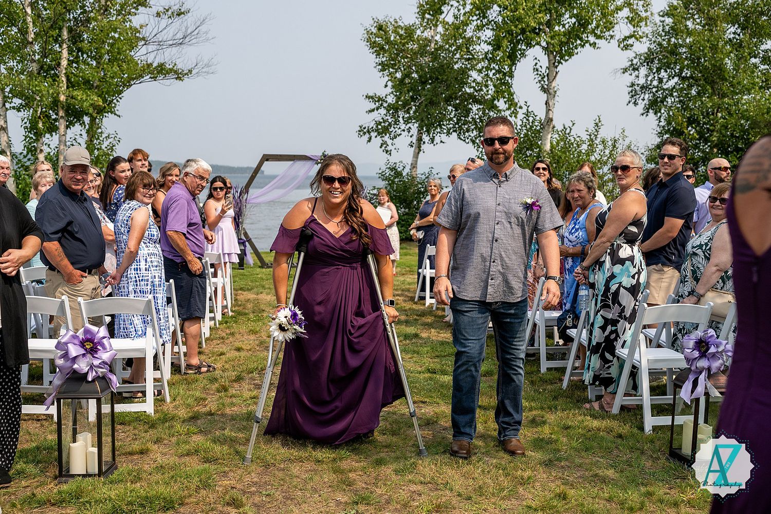 Wedding party members dance joyfully down the aisle between white chairs at an outdoor ceremony with trees in background.