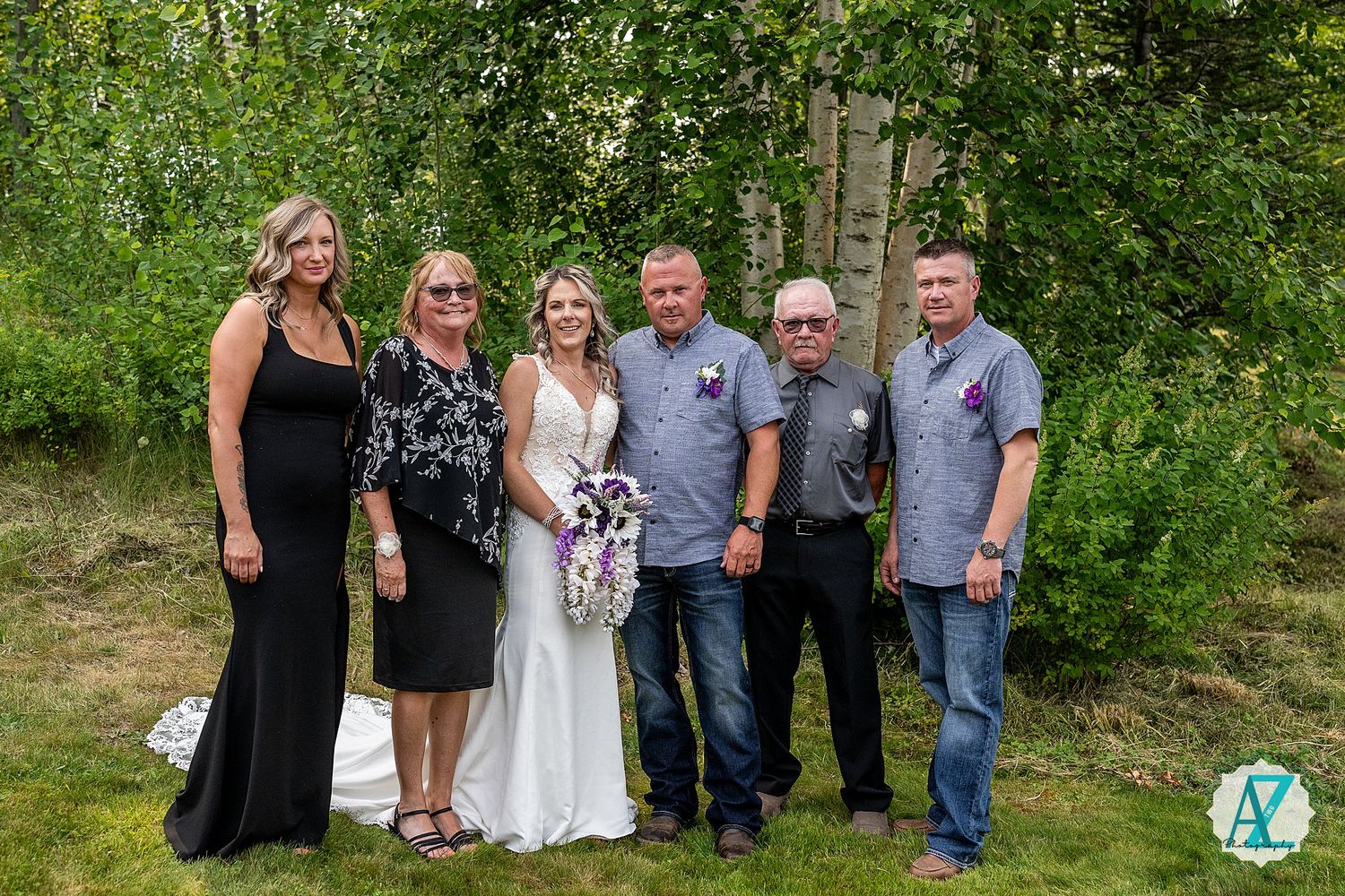 A group of people standing together outdoors on a sunny day with trees and greenery in the background.