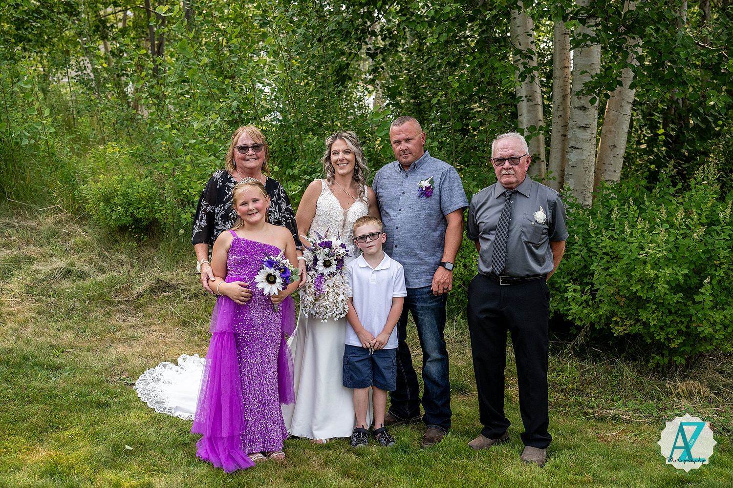 A group of six people in dressy attire pose together outdoors against a green natural backdrop.