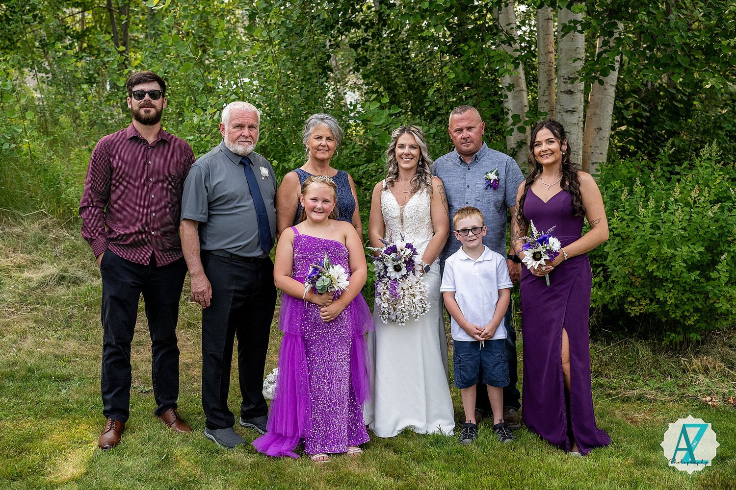 A wedding party poses outdoors in the summer with greenery and trees in the background, featuring purple bridesmaids dresses.