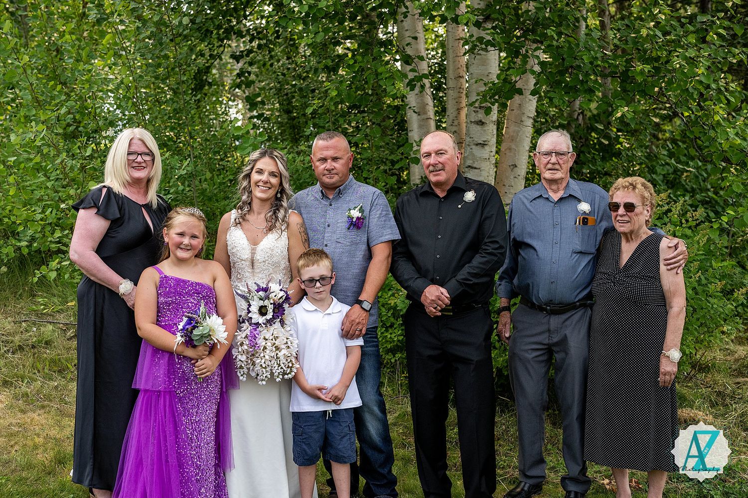 A wedding party group poses together outdoors against a green leafy background in summer attire.