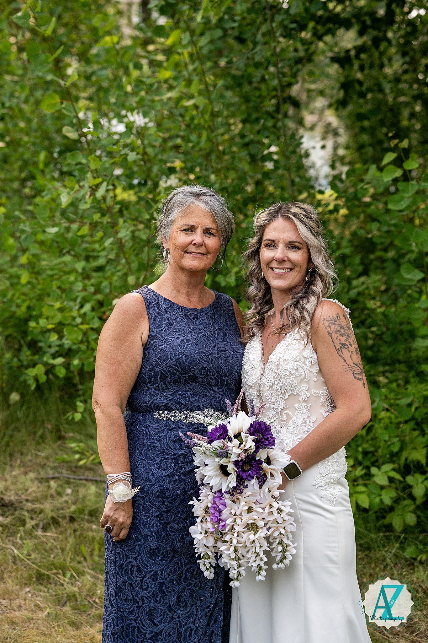 Two people in navy blue and white dresses pose together outdoors holding a purple and white floral bouquet.