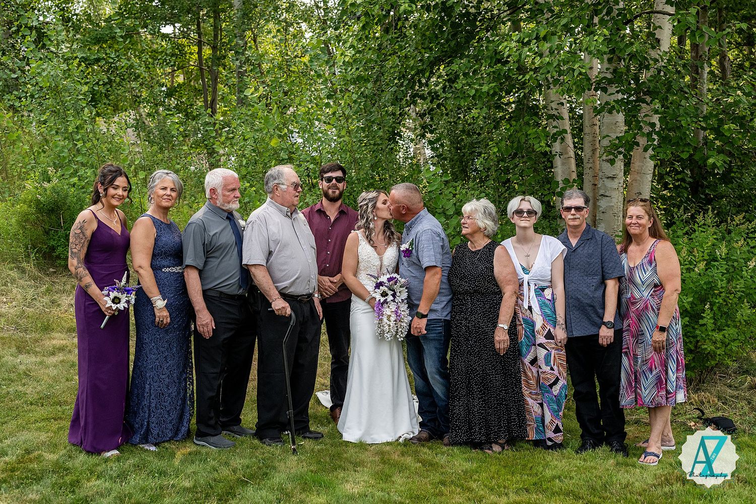Wedding group photo with guests in various dresses and attire gathered outdoors against a green wooded backdrop.