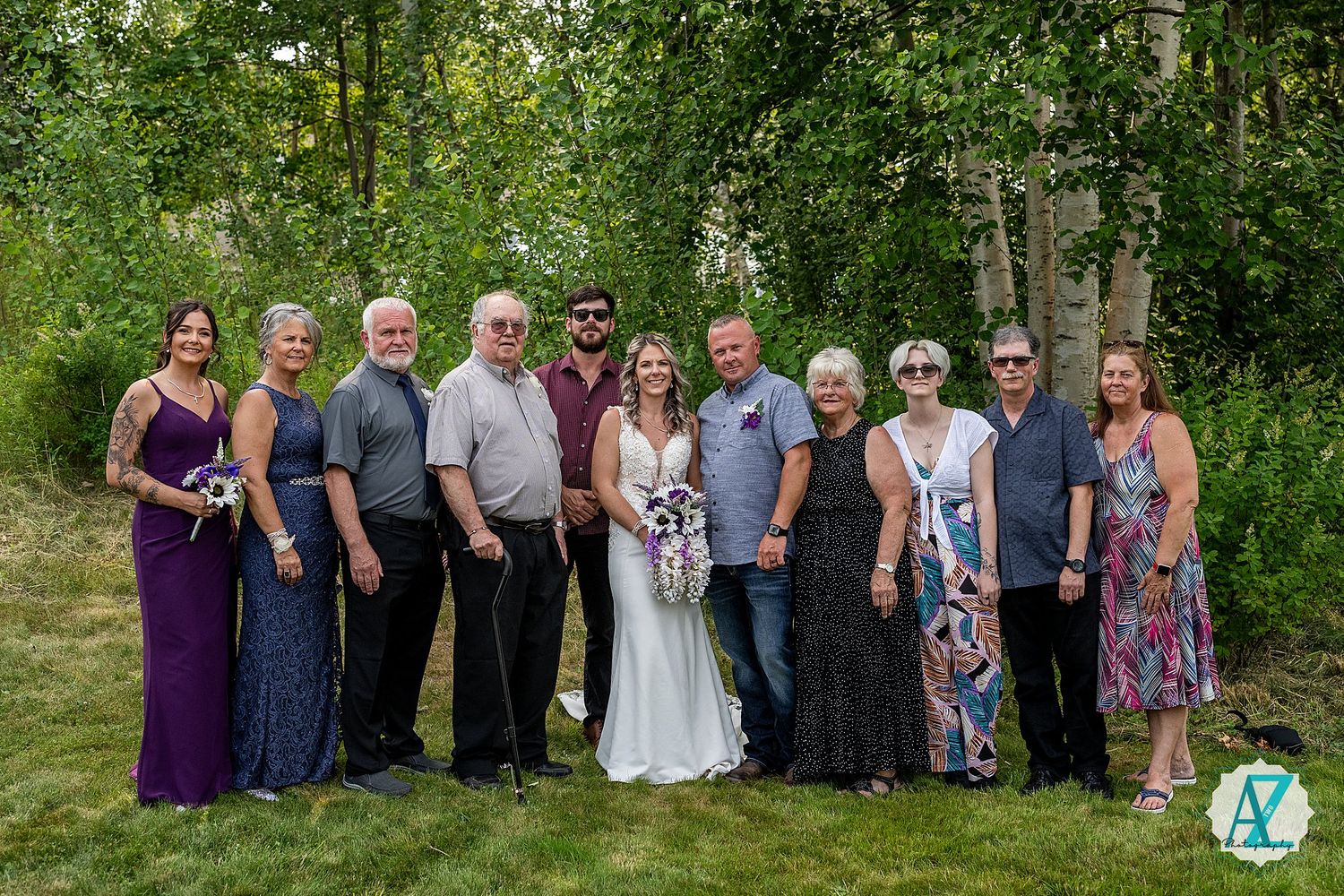 Purple and gray wedding party poses together outdoors in front of trees and lush greenery.