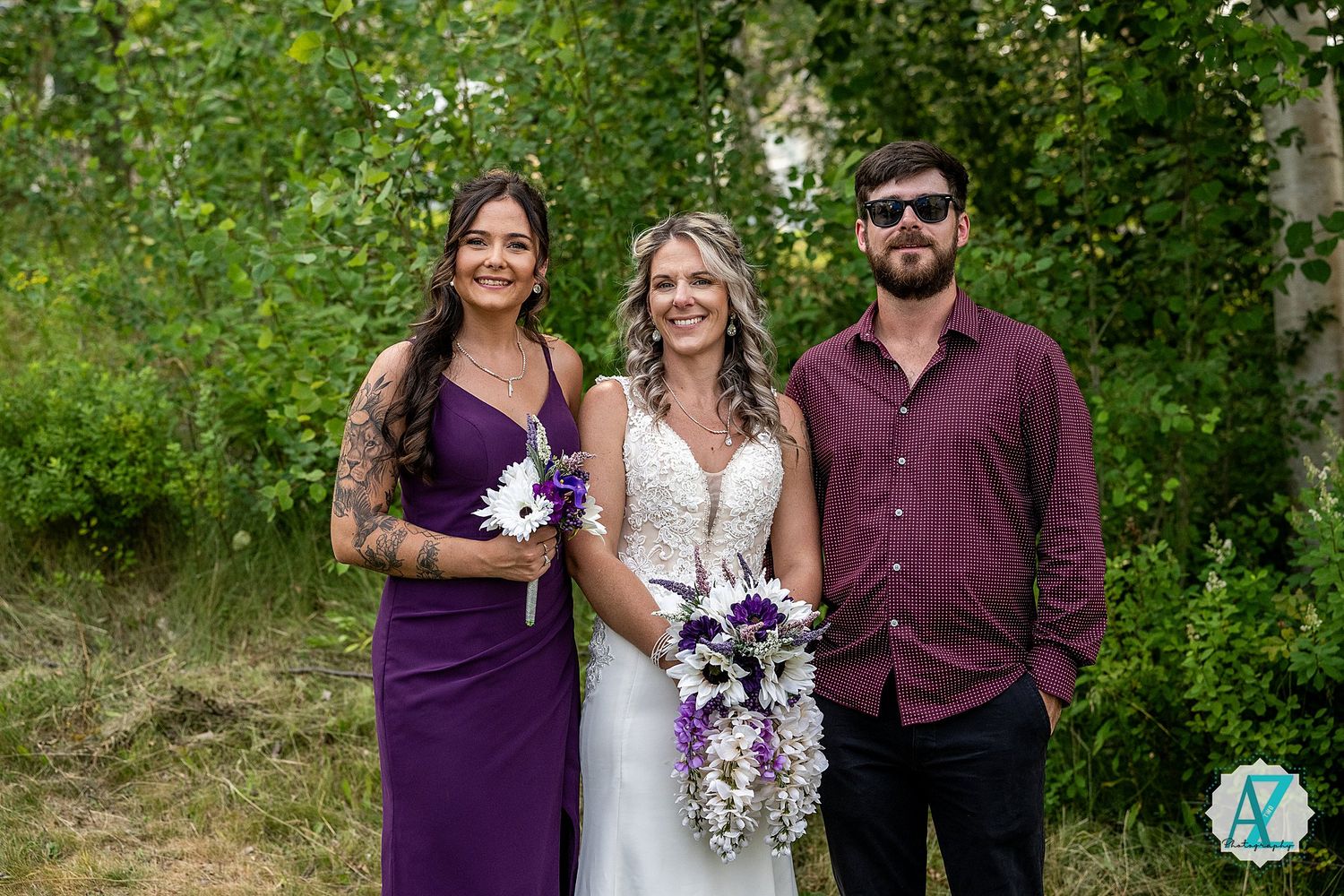 Three people in wedding attire pose together outdoors, featuring a purple dress and burgundy shirt.
