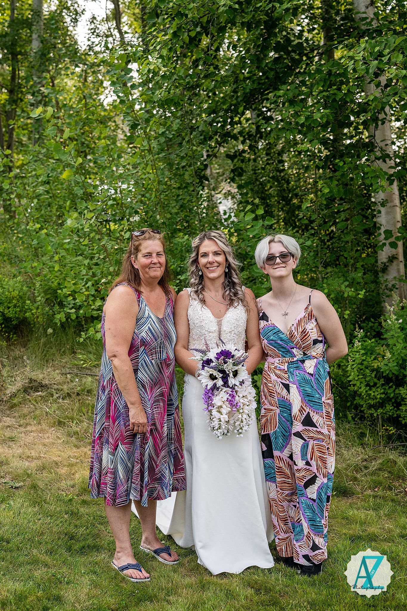 Three people in colorful summer dresses pose together outdoors with greenery and trees in the background.