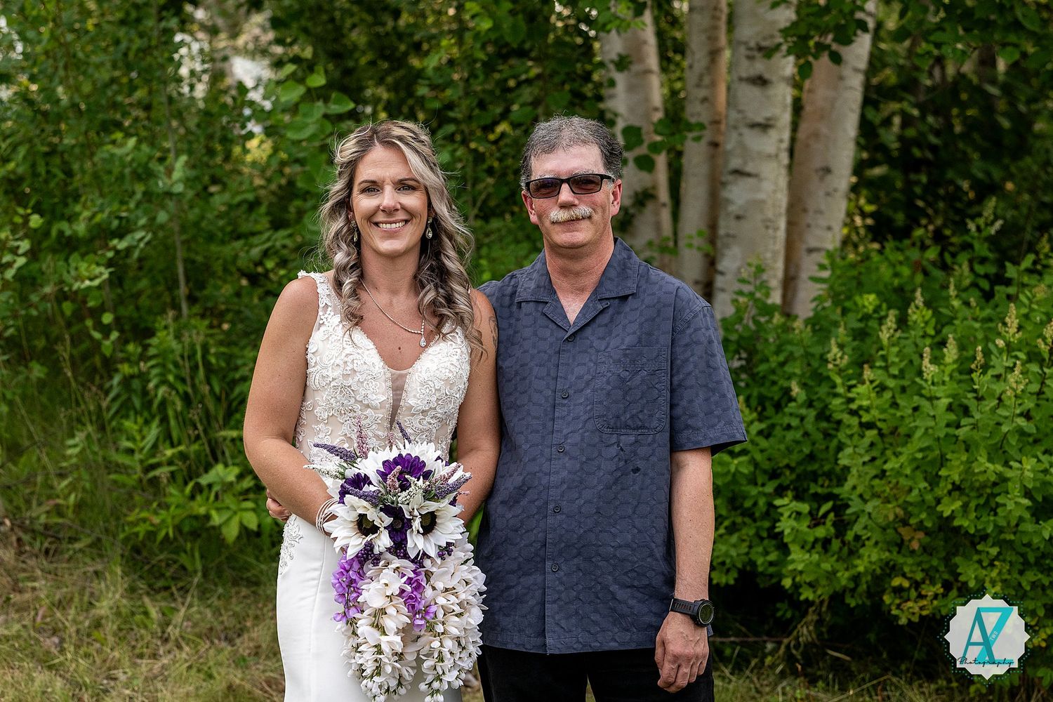 Couple stands together in a lush green forest setting with natural sunlight filtering through the trees.