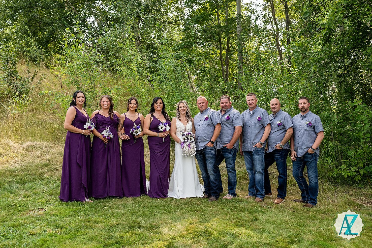 Wedding party in formal attire poses outdoors against lush green trees and foliage.