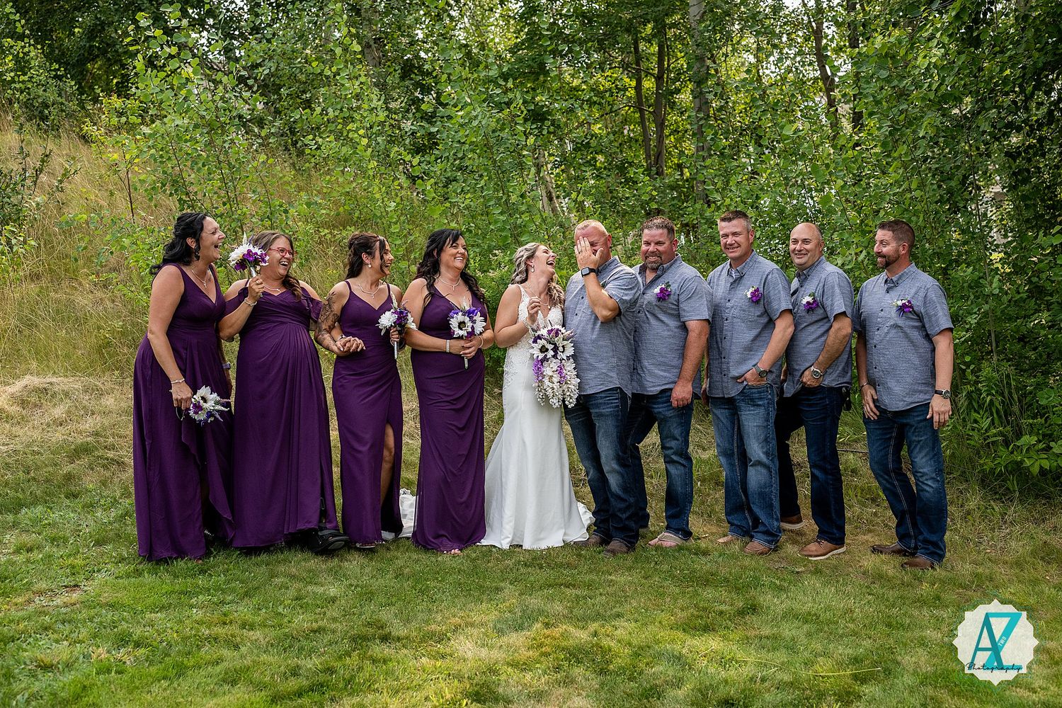 A wedding party poses outdoors in a line surrounded by lush green trees and natural scenery.