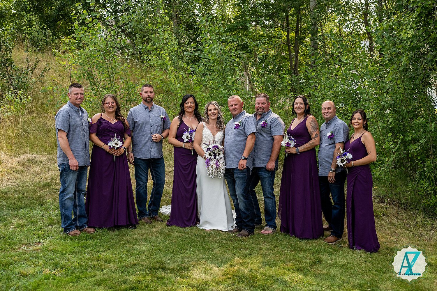 Wedding party in purple dresses poses outdoors against lush green foliage backdrop during summer celebration.