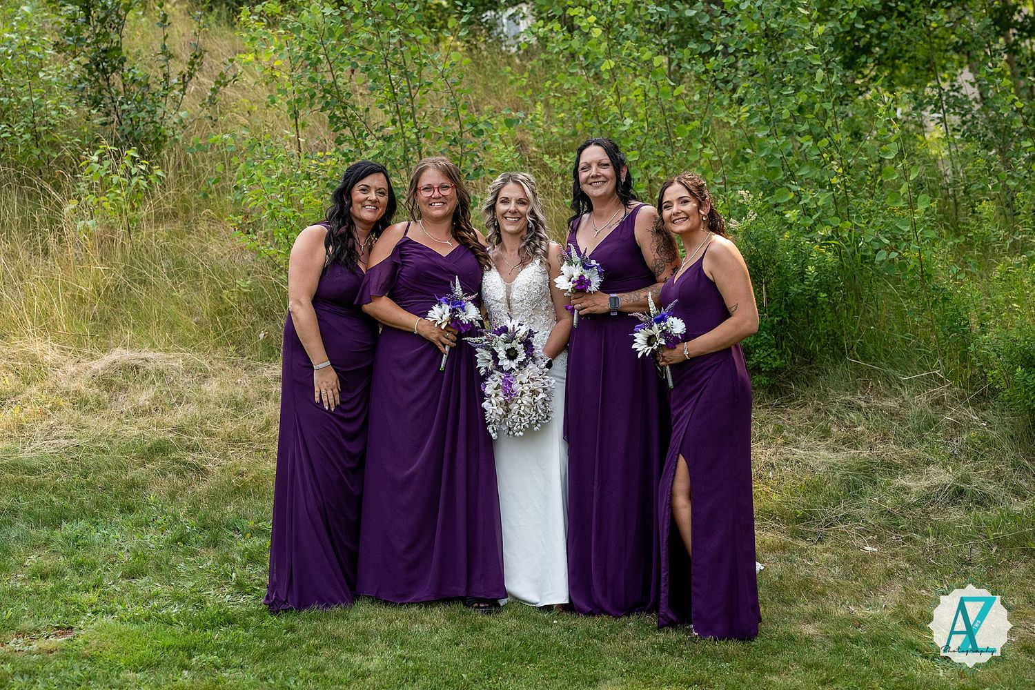 A group in matching deep purple bridesmaid gowns and one in white stand together outdoors against greenery.