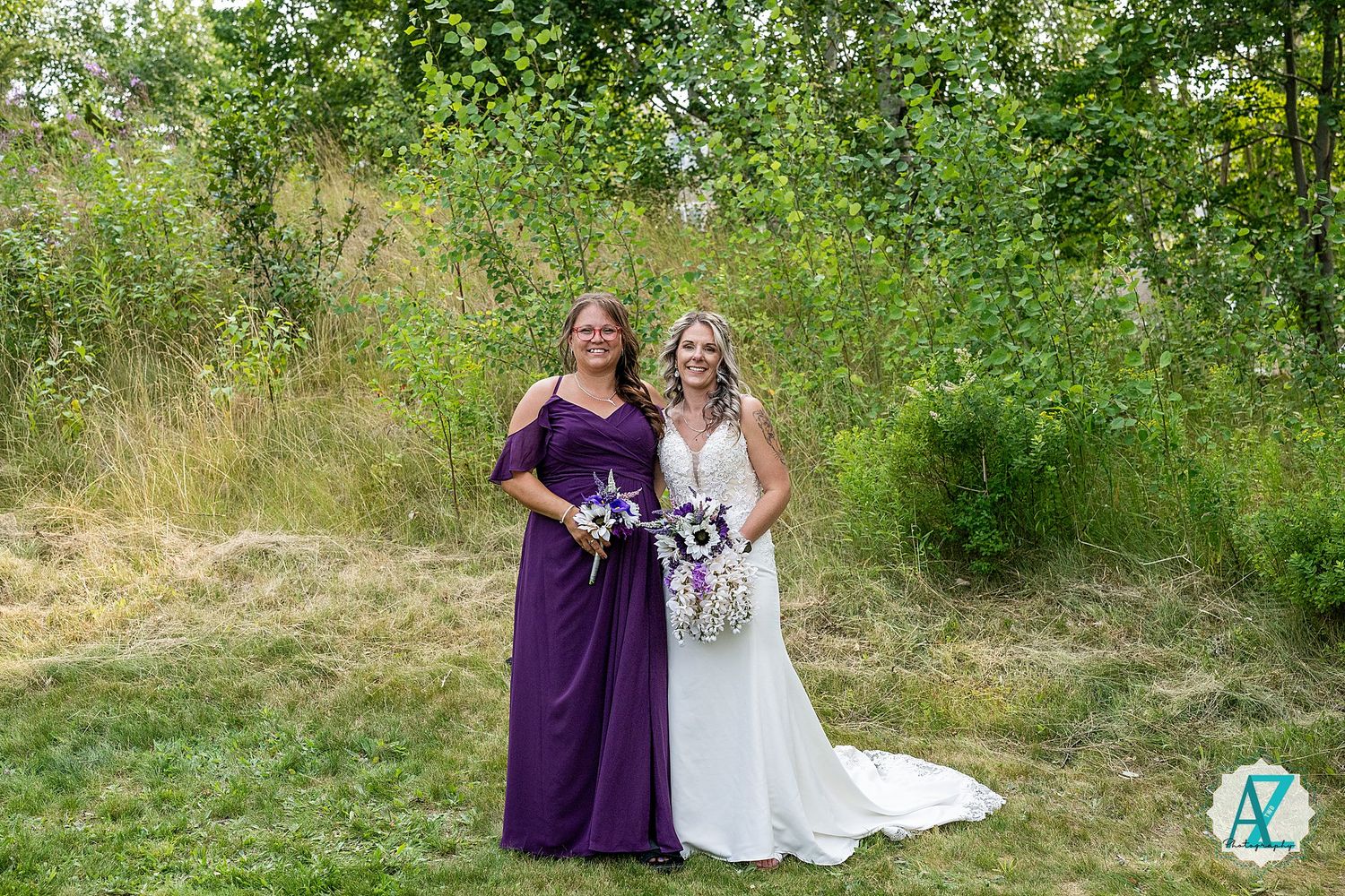 Two people stand together outdoors in formal dresses holding bouquets among greenery and tall grass.