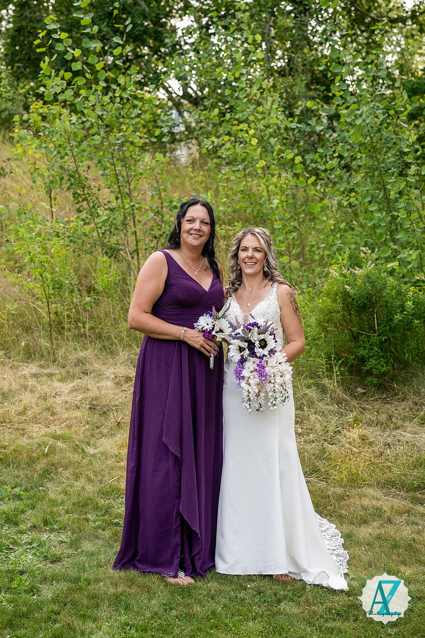 Two women pose together outdoors at a wedding, wearing a purple bridesmaid dress and white bridal gown with matching bouquets.