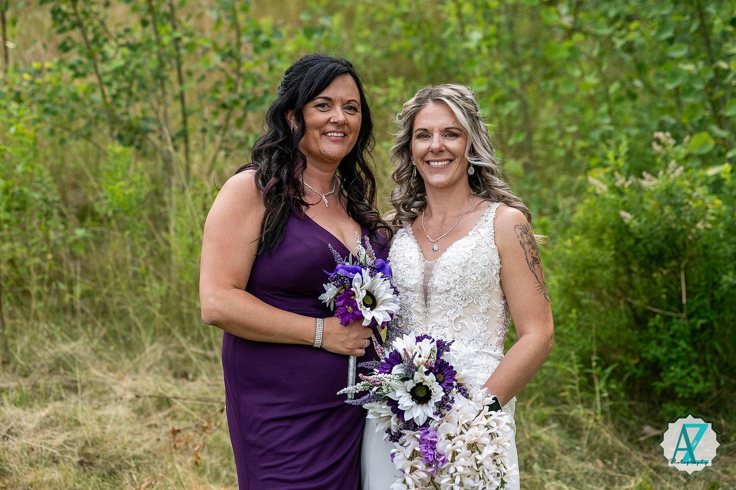 Two people pose together outdoors in purple and white attire during a wedding celebration with natural greenery behind them.