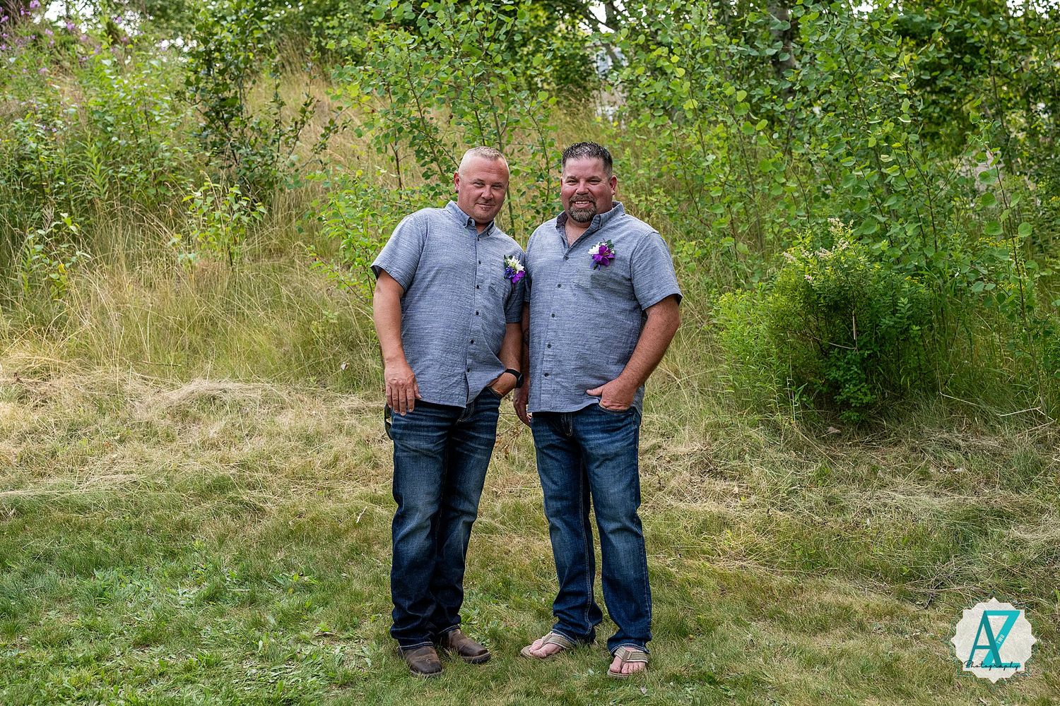 Two people stand together on a grassy hillside surrounded by trees and greenery in an outdoor setting.