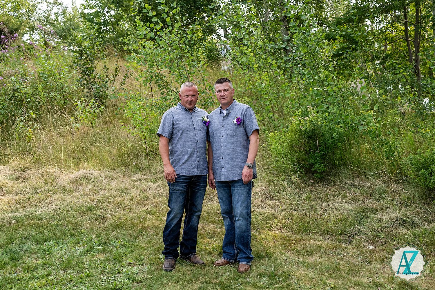 Two people wearing matching gray shirts stand together on grassy terrain with green foliage in the background.