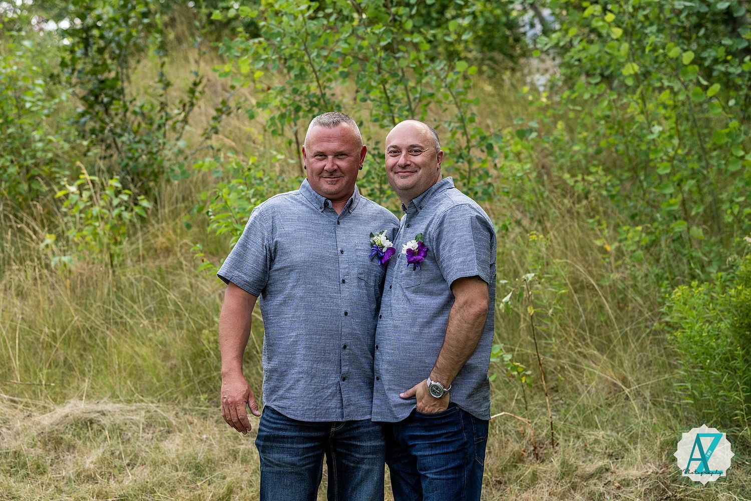 Two people wearing casual attire stand together smiling outdoors with green foliage in the background.