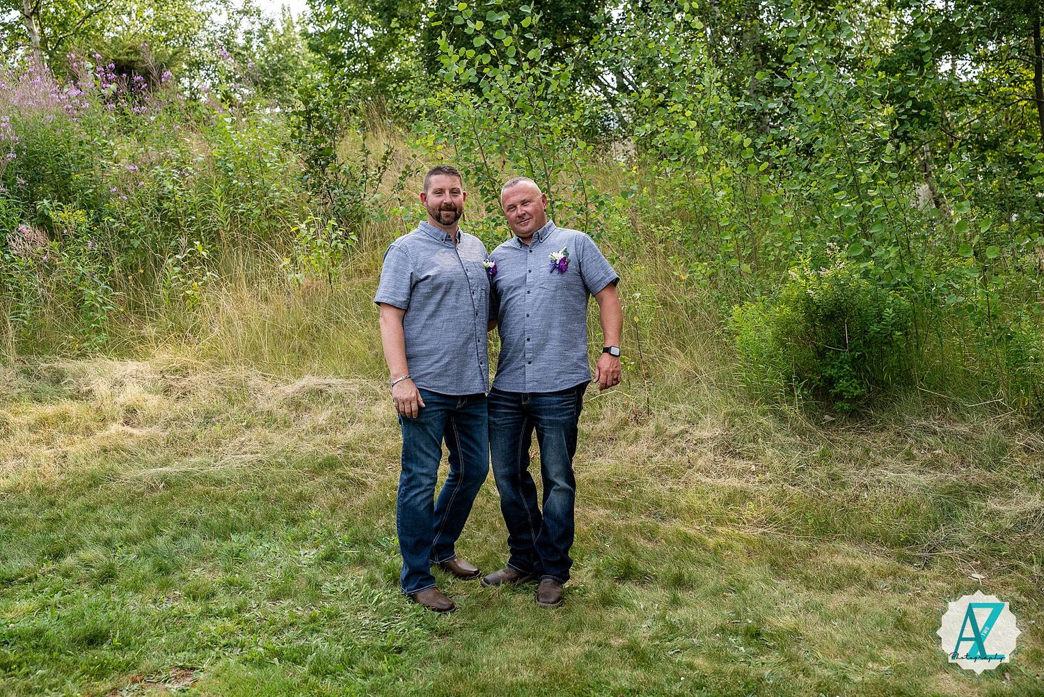 Two people in matching gray shirts stand together outdoors on grassy ground with lush greenery in the background.