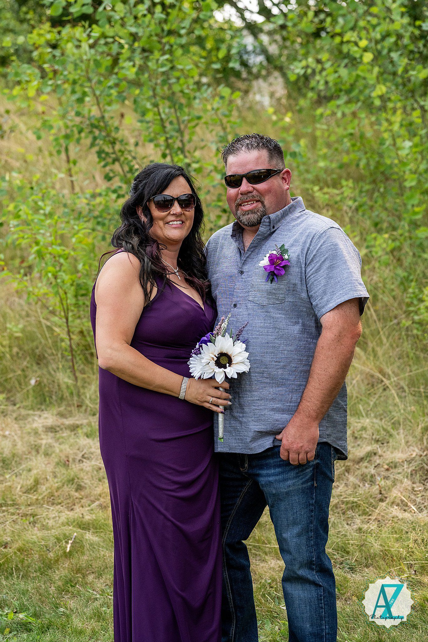 A couple stands together outdoors in formal attire, with one in a purple dress holding white flowers and one in grey.