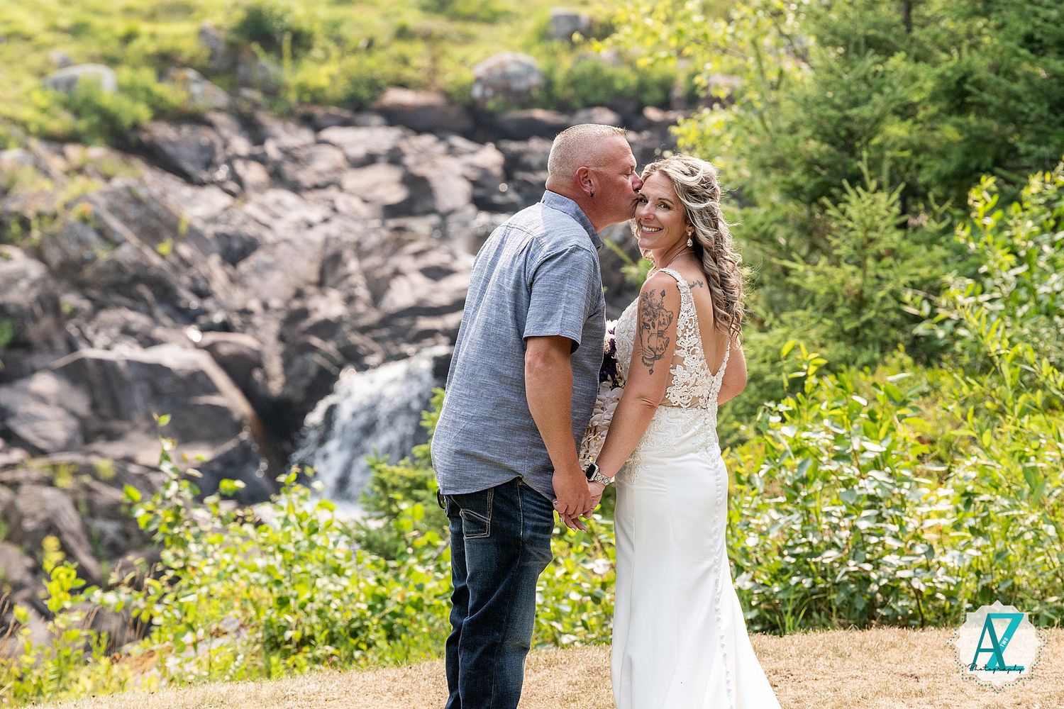 Engaged couple sharing romantic moments along a rocky coastal shoreline surrounded by lush greenery in summer.