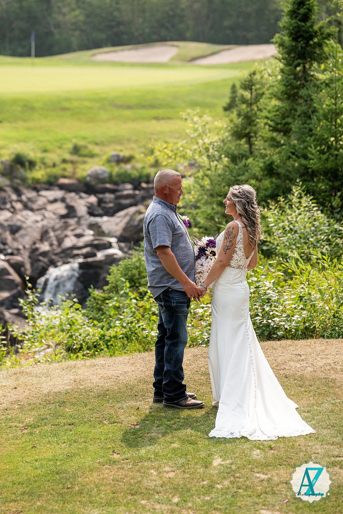 A couple stands together on a grassy hill overlooking a rocky waterfall on their wedding day.