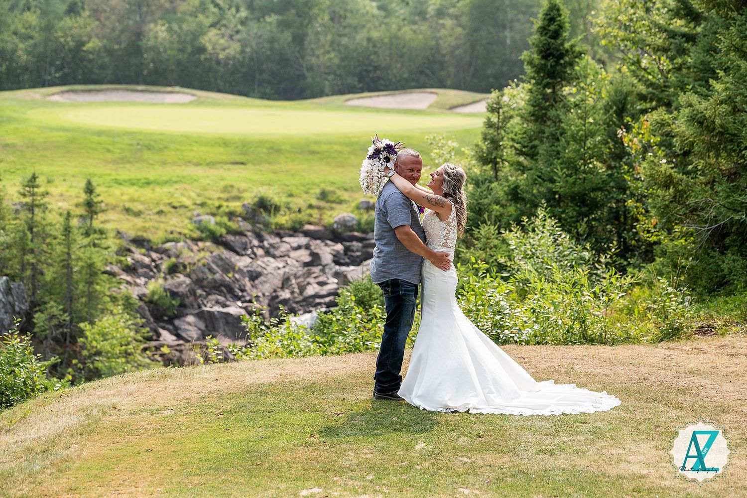 A couple embraces on a grassy cliffside overlooking evergreen trees and rocks at a scenic golf course venue.
