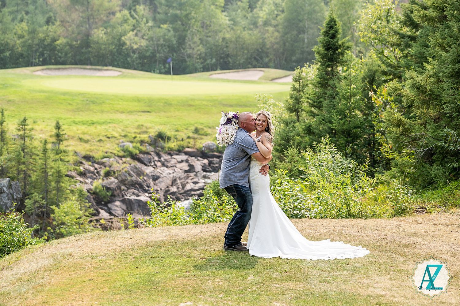 Two people in wedding attire embrace against a scenic mountain landscape backdrop with lush evergreen trees.