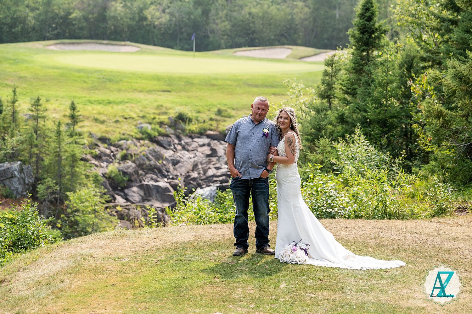 A couple stands together on a hilltop overlooking a scenic golf course surrounded by evergreen trees and rocky terrain.