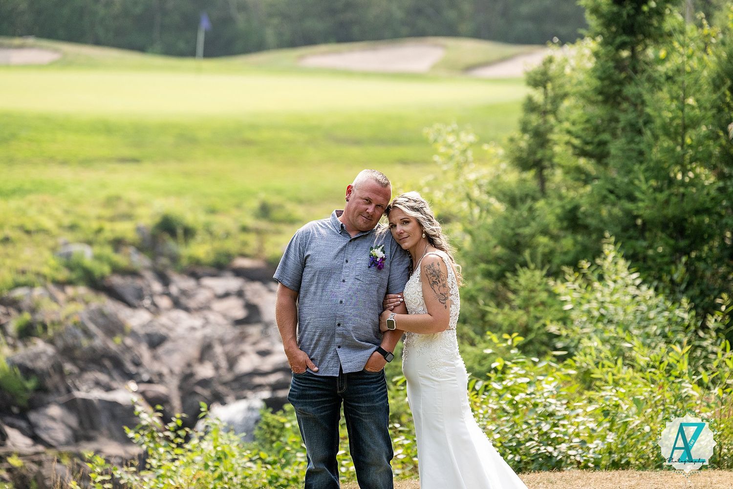 A couple stands together outdoors near rocky terrain with lush green fields and trees in the background.