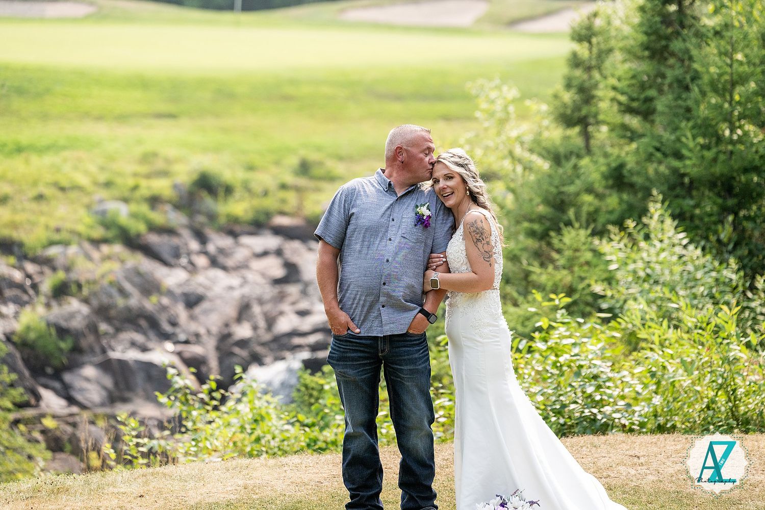 A couple shares an intimate moment on a scenic mountain trail with rocky terrain and lush greenery in the background.